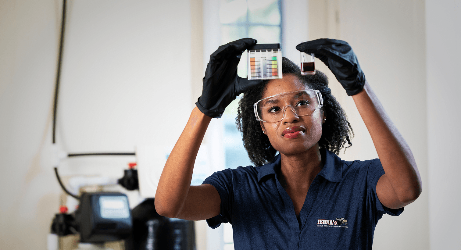 A IAERNA's technician in a blue uniform and safety gear compares a water sample to a testing chart in front of a water softener system.