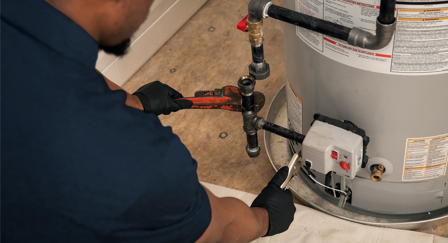 An IERNA'S plumber wearing black gloves uses a pipe wrench to repair the gas line and control valve assembly on a residential water heater tank.