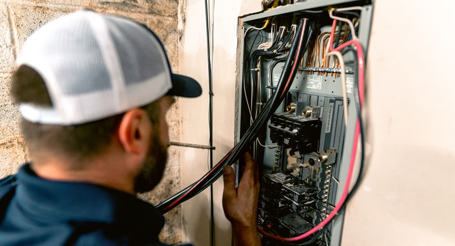 A Sansone electrician in a baseball cap inspects and installs thick black and red electrical wires inside an open residential circuit breaker panel box on a basement wall.